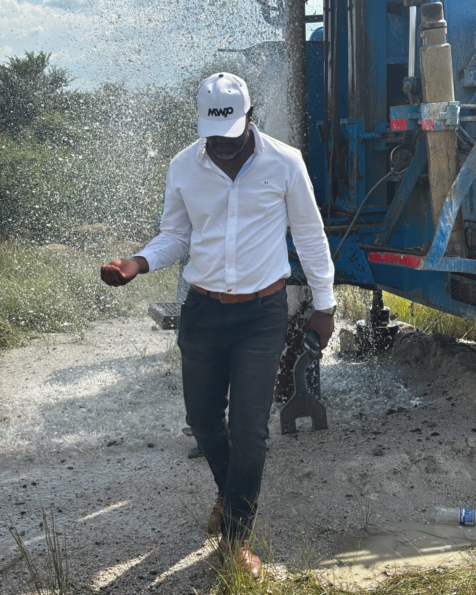Antony Dube in a white cap and shirt at a borehole wellhead, water spraying behind him